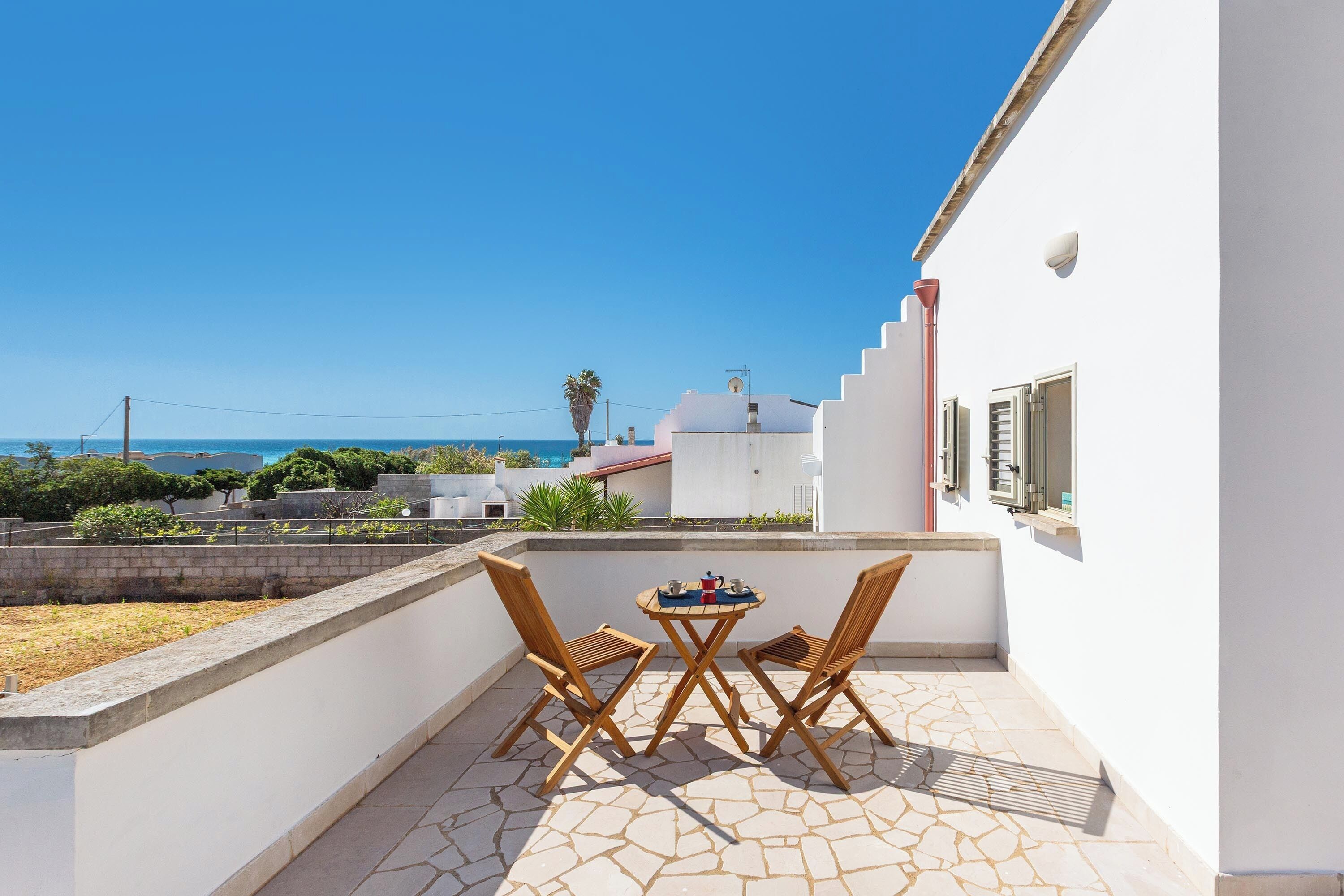 A balcony with two wooden chairs and a small table, overlooking a coastal area with a clear sky.