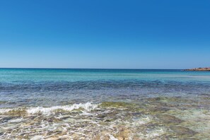 A clear beach with shallow waters revealing rocks and pebbles, a clear sky, and a distant shoreline with buildings. - Domus Maris Felloniche by BarbaHouse (Castrignano del Capo)