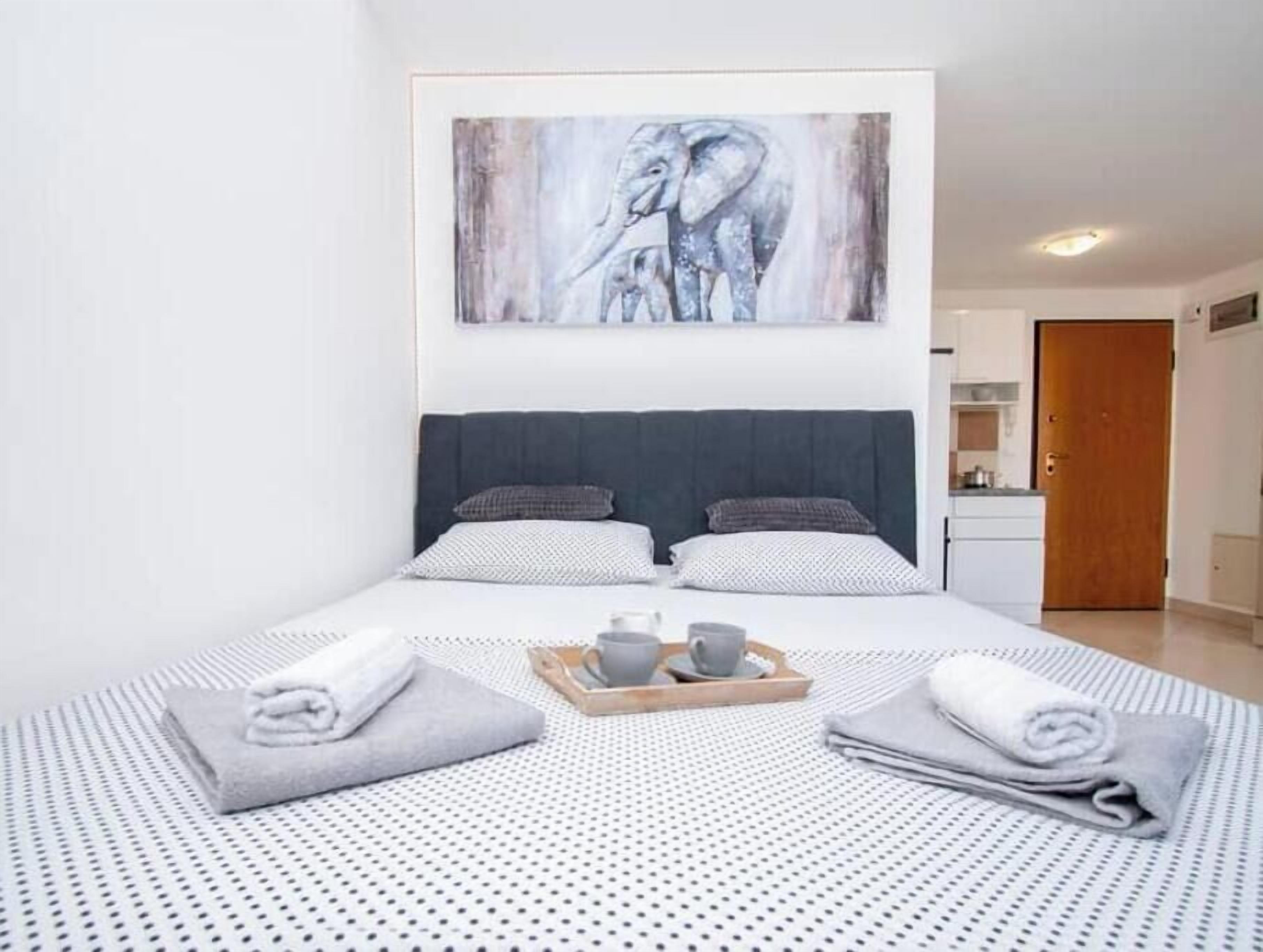 A hotel room with a large bed, a painting of an elephant, a small kitchenette, and a wooden door.