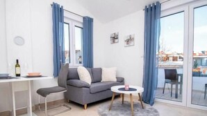 Een moderne woonkamer met een grijze bank, een klein rond tafeltje, een eettafel met stoelen, en een glazen deur naar een buitenruimte.