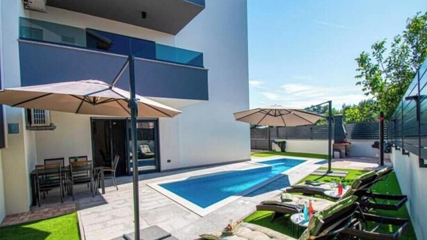 Un área de piscina moderna al aire libre con sillones de descanso y sombrillas, rodeada de un césped cuidado y un edificio con techo plano.