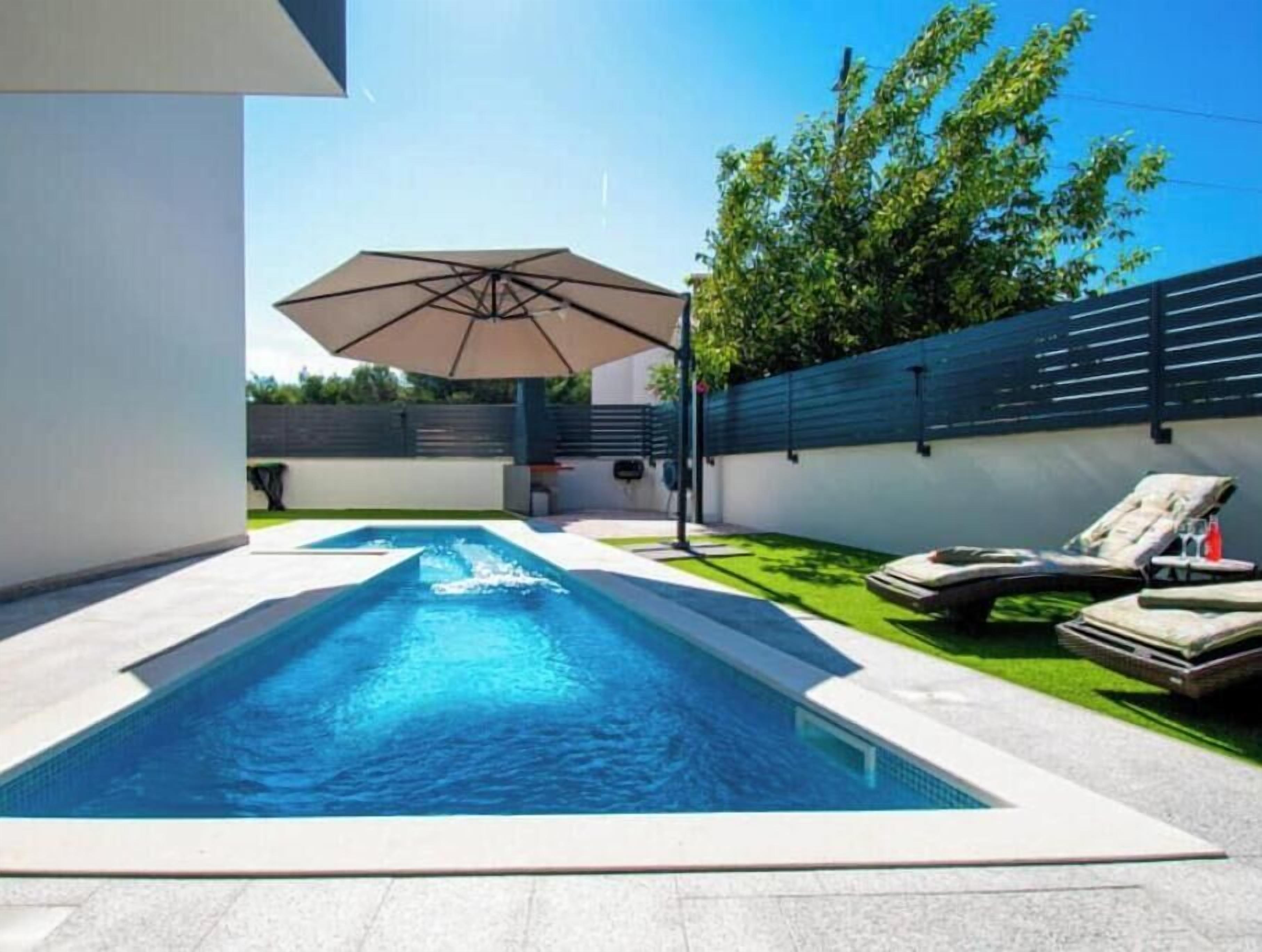Une piscine avec un espace détente, une pergola et une pelouse verdoyante.