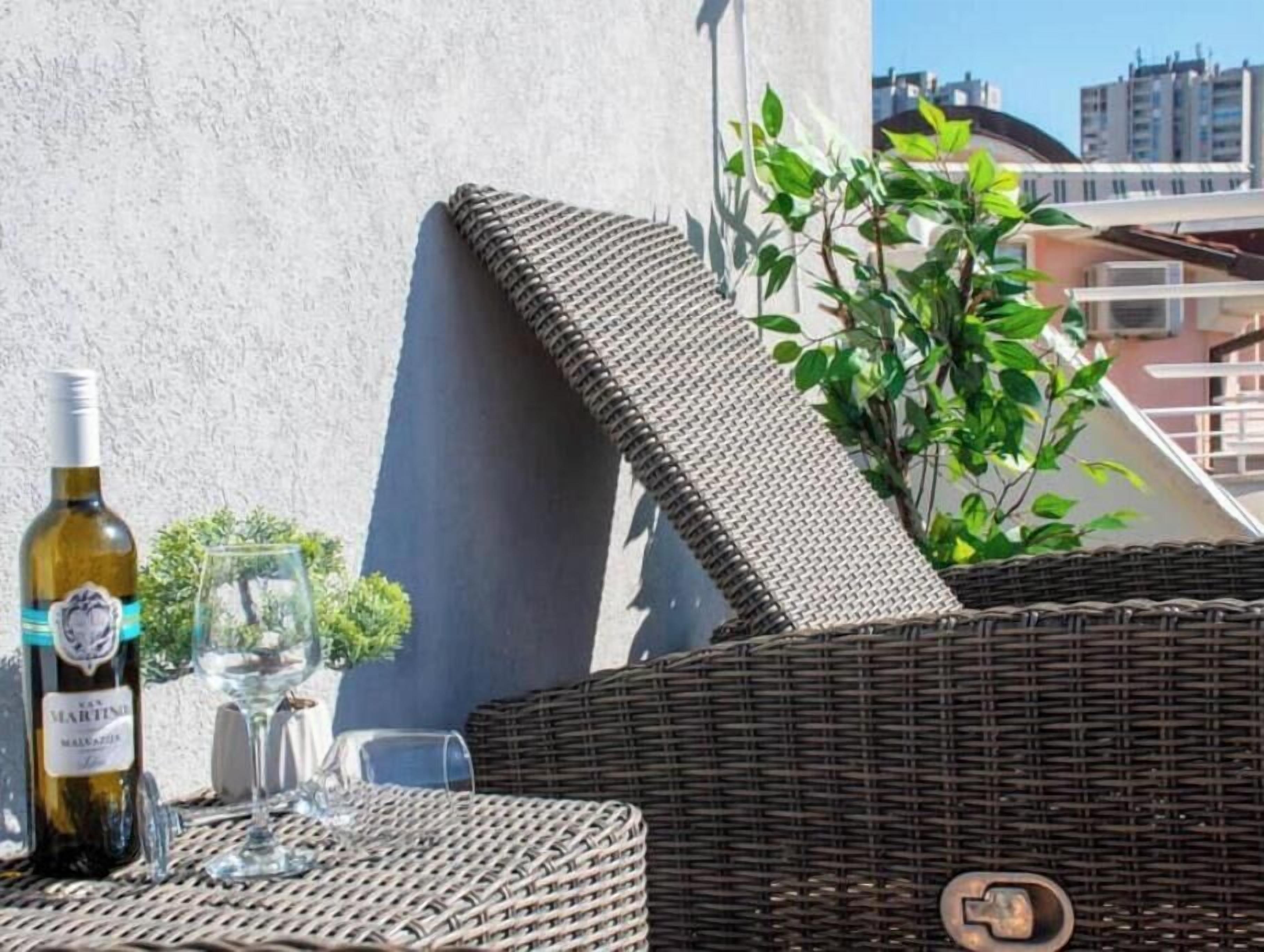 A wicker outdoor seating area with a bottle of wine and glasses, a potted plant, and a view of buildings in the background.