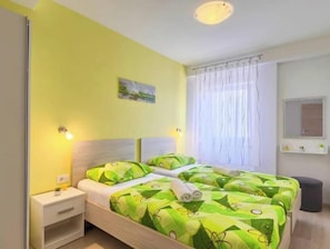 Ein Doppelbett mit grün gemusterten Betttextilien, ein Nachttisch, ein Fenster mit weißen Vorhängen und ein kleiner Spiegel an der Wand.