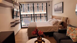 Una camera d'hotel moderna con un letto, una poltrona, una TV, una finestra con persiane e vista sulla città.