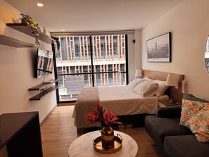 Ein modernes Hotelzimmer mit Bett, einer Sitzecke, einem Fernseher, einem Fenster mit Jalousien und Blick auf die Stadt.