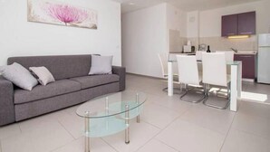A modern living room with a glass coffee table, grey sofa, and a dining area with white chairs. - A01 - C: Lotus Apartment (Marčana)