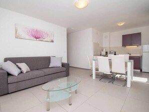 A modern living room with a glass coffee table, grey sofa, and a dining area with white chairs. - A01 - C: Lotus Apartment (Marčana)