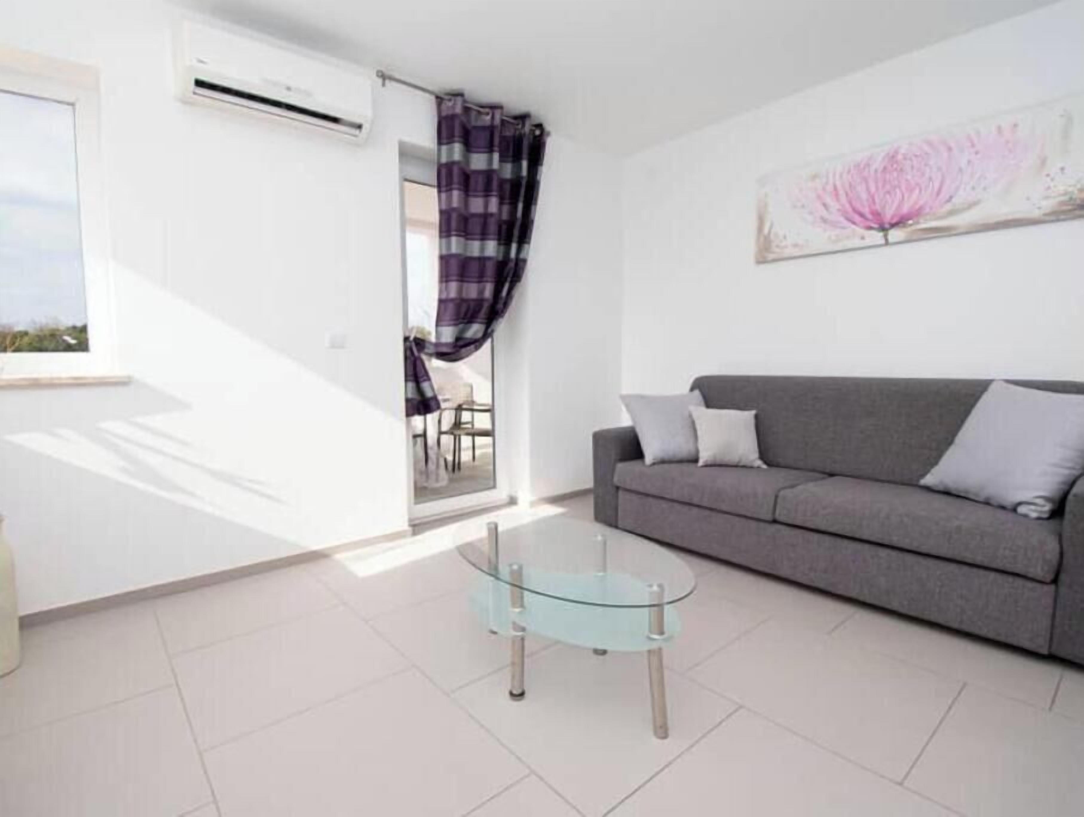 A modern living room with a glass coffee table, a grey sofa, and a large window with curtains.