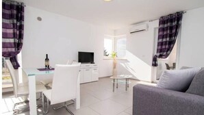 A modern living room with a grey sofa, a dining area with a white table and chairs, and a television. - A01 - C: Lotus Apartment (Marčana)