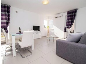 A modern living room with a grey sofa, a dining area with a white table and chairs, and a television. - A01 - C: Lotus Apartment (Marčana)