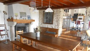 Un comedor con una gran mesa de madera y bancos, una chimenea de piedra y una cocina al fondo.
