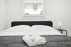 Un lit bien fait, avec du linge de lit blanc, une couverture grise à texture et une petite table de chevet surmontée d’une lampe.