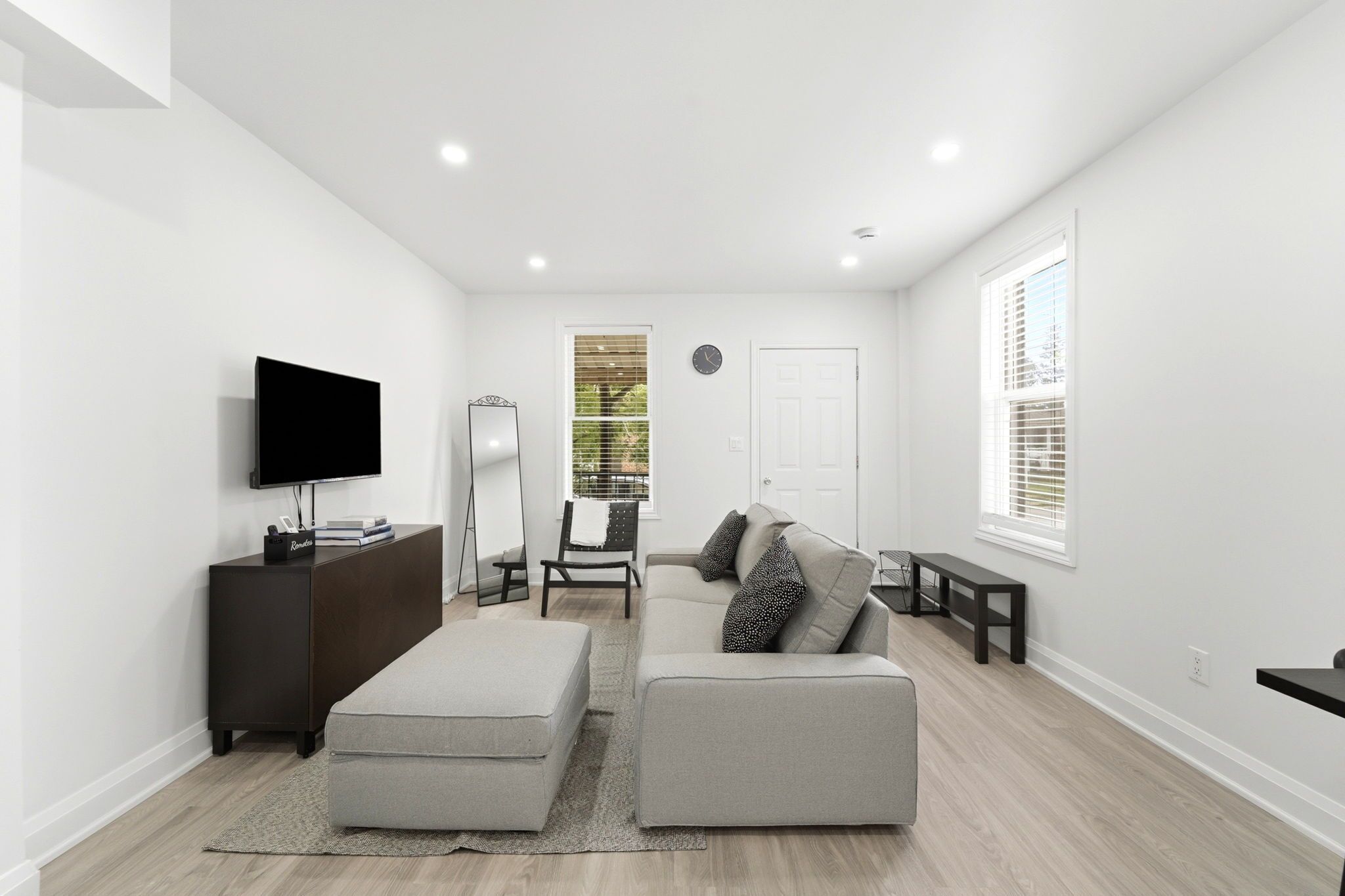 Een moderne woonkamer met een flatscreen televisie, een donker houten kast, een grijze bank met gekleurde kussens, een witte deur en een raam met zonwering.