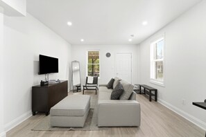 Un salon moderne avec un téléviseur à écran plat, un meuble en bois foncé, un canapé gris agrémenté de coussins à motifs, une porte blanche et une fenêtre avec des stores.