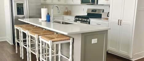 A kitchen with white cabinets, a stainless steel refrigerator, a microwave, a stove, and a bar stool seating area.
