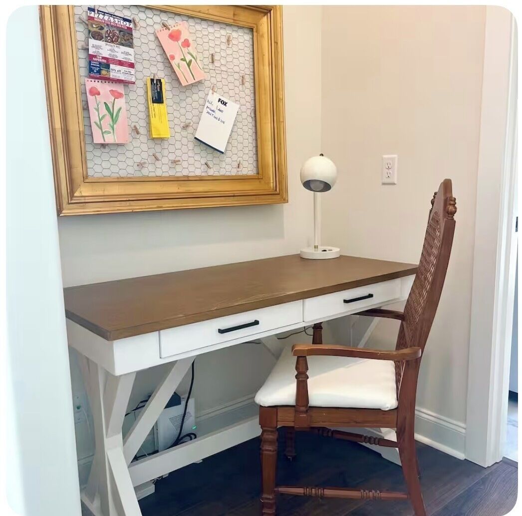 A wooden desk with a white drawer, a framed bulletin board with notes, a lamp, and a wooden chair.