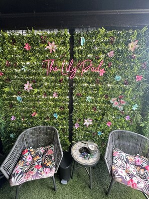 Un patio con dos sillas de mimbre, una mesa redonda pequeña y una pared decorativa con flores colgantes.