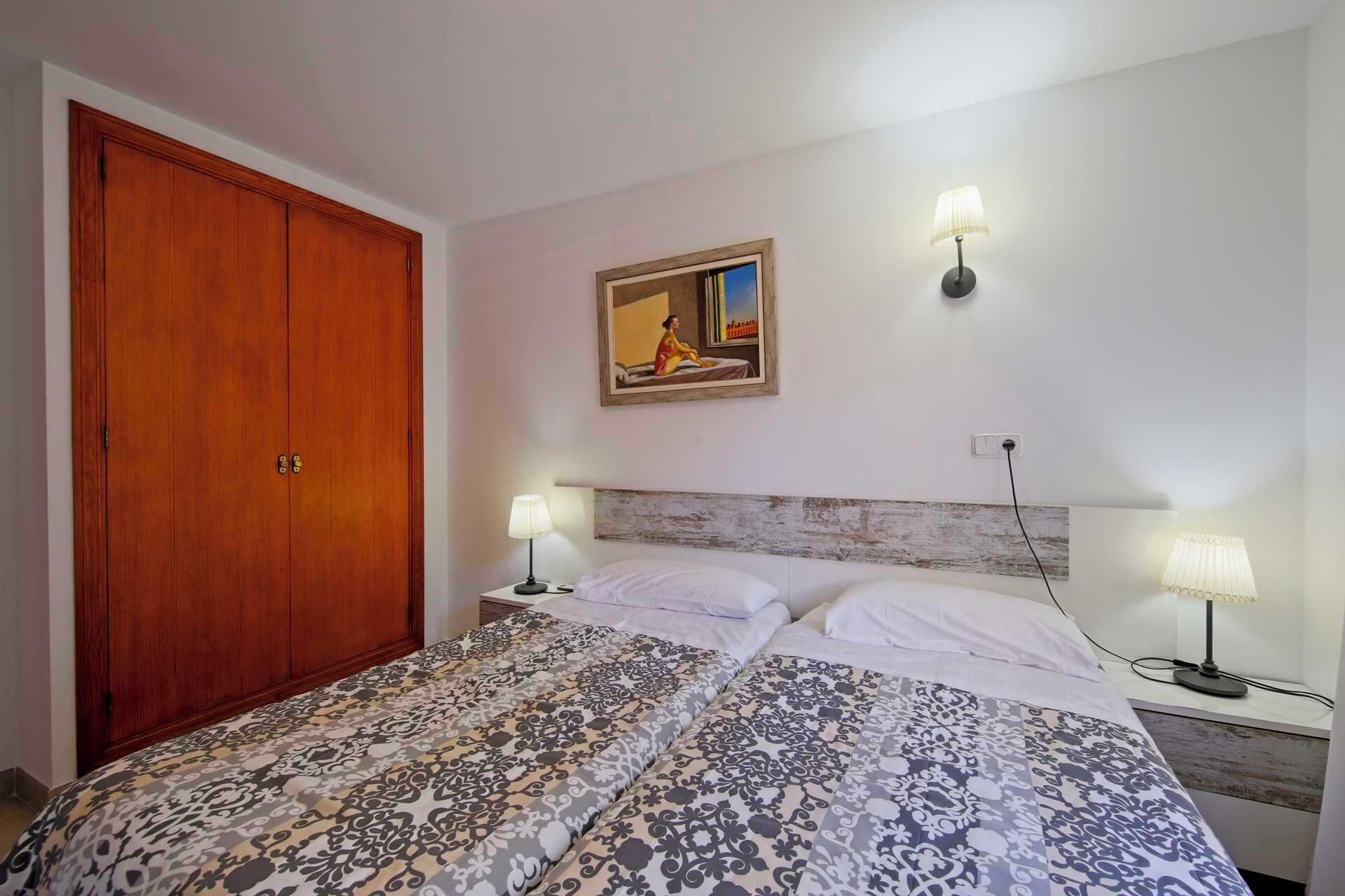 A bedroom with a wooden wardrobe, a bed with patterned bedding, two bedside lamps, and a painting on the wall.