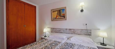 A bedroom with a wooden wardrobe, a bed with patterned bedding, two bedside lamps, and a painting on the wall.