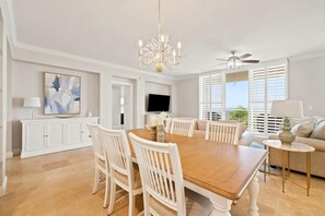 A dining area with a wooden table and white chairs, a chandelier, a ceiling fan, and a television on a cabinet. - Indigo East 402 (Pensacola)