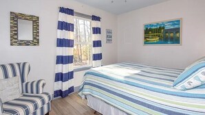 Un dormitorio con cama, una silla, ventilador de techo, ventana con cortinas y un cuadro en la pared.