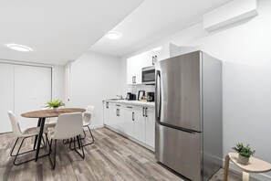 Eine moderne Küche mit weißen Schränken, einem Edelstahlkühlschrank, einer Mikrowelle und einem Essbereich mit einem Holztisch und weißen Stühlen.