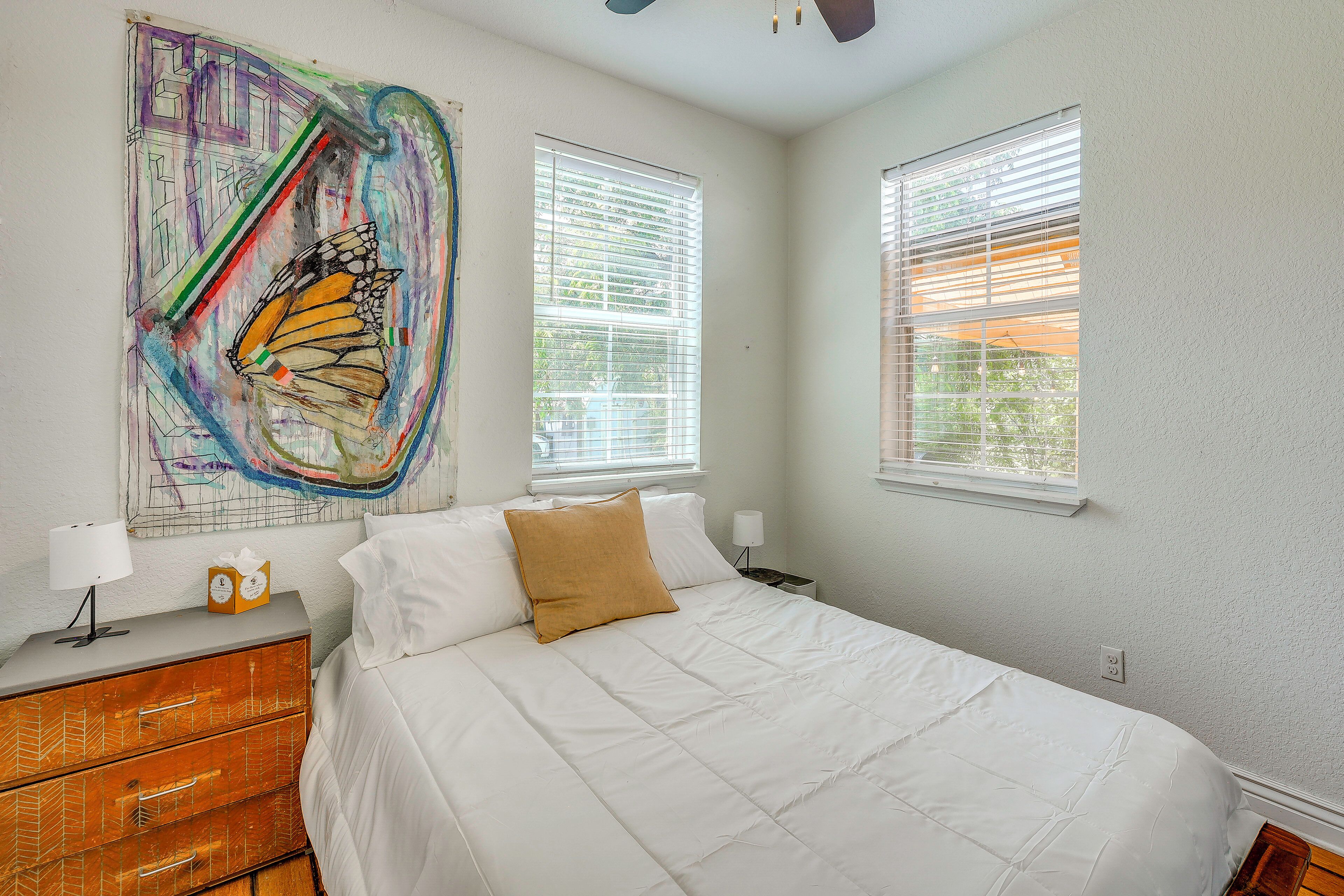 A bedroom with a bed, a dresser, a painting, and two windows with blinds.