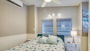 Quarto com uma cama, mesa de cabeceira, luminária, ventilador de teto e duas janelas com cortinas.