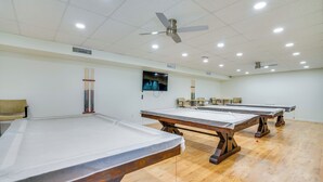Um espaço com várias mesas de bilhar, ventoinhas no teto, iluminação embutida e uma televisão na parede.