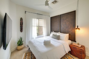 A modern bedroom with a bed, a ceiling fan, a window with blinds, a wall-mounted TV, and a decorative plant.
