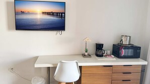 Een minimalistisch ingerichte kamer met een wit bureau, een witte stoel, een koffiezetapparaat, een magnetron en een televisie waarop een zonsondergang te zien is.