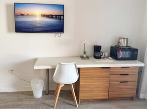 Ein schlicht eingerichteter Raum mit weißem Schreibtisch, weißem Stuhl, einer Kaffeemaschine, einer Mikrowelle und einem Fernseher, der eine Sonnenuntergangsszene zeigt.