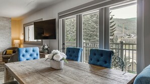 Uma sala de jantar com uma mesa de madeira, cadeiras estofadas em azul, um vaso de flores e uma televisão fixada na parede.
