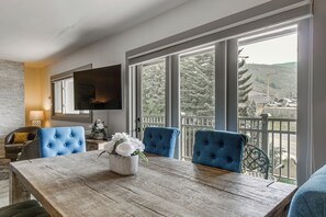 Ein Essbereich mit einem Holztisch, blau gepolsterten Stühlen, einer Blumenvase und einem an der Wand befestigten Fernseher.