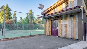 Ein hölzernes Gebäude mit einem Schild, auf dem 'The CLUBHOUSE' steht, und ein eingezäunter Tennisplatz im Hintergrund.