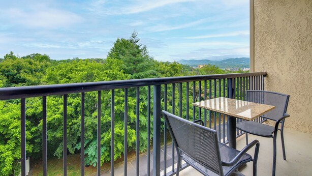 A balcony with a table and chairs, overlooking a lush green landscape and distant mountains. - Mtn Views, Pool Access! Walkable Gem Near Parkway (Pigeon Forge)