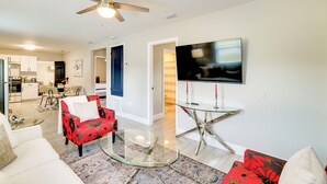 A modern living room with a glass coffee table, a red floral sofa, a flat-screen TV mounted on the wall, and a ceiling fan. - Modern Lakeland Retreat w/ Office I Fenced Yard (Lakeland)