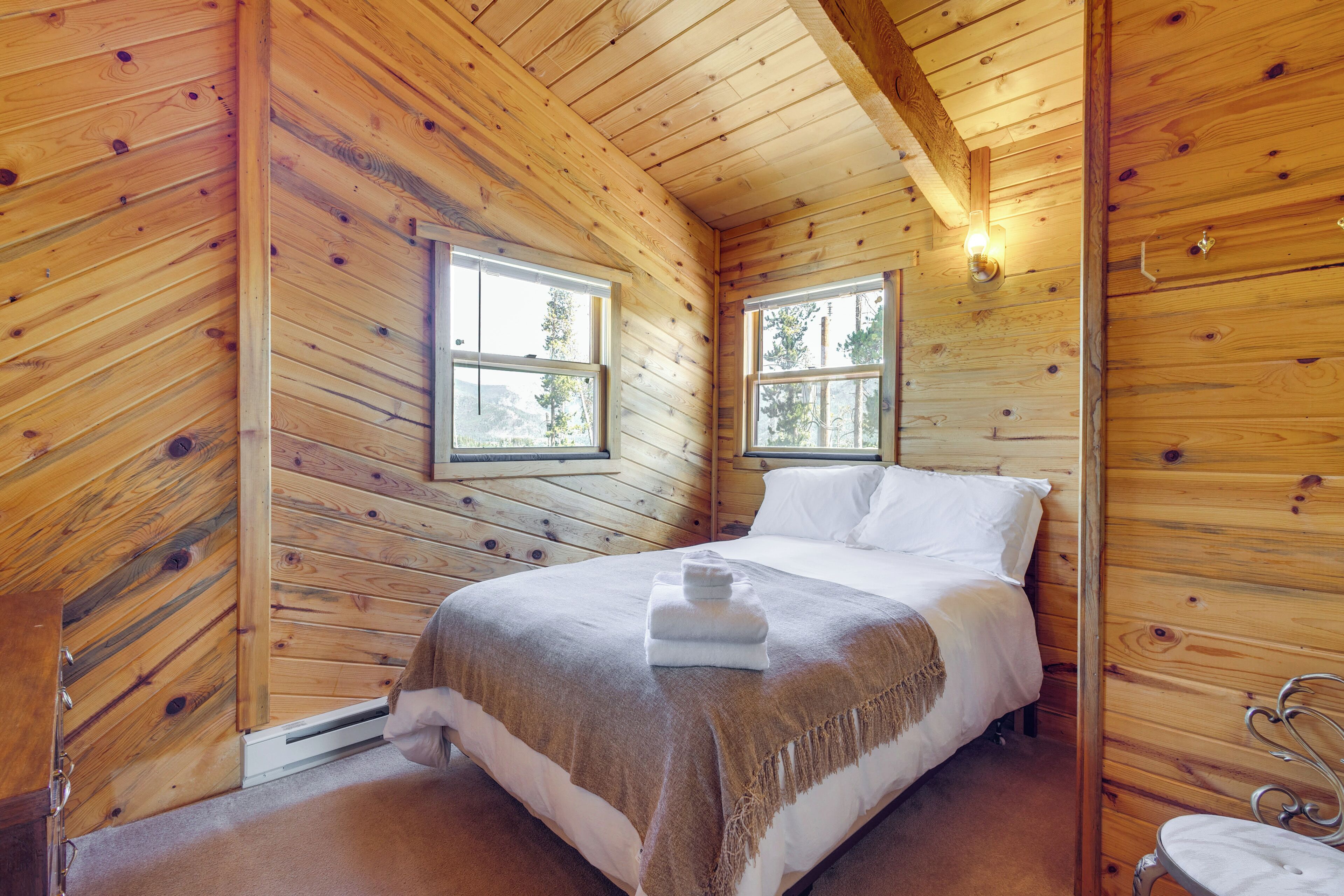 A wooden cabin bedroom with a bed, two windows, and a wall-mounted light fixture.