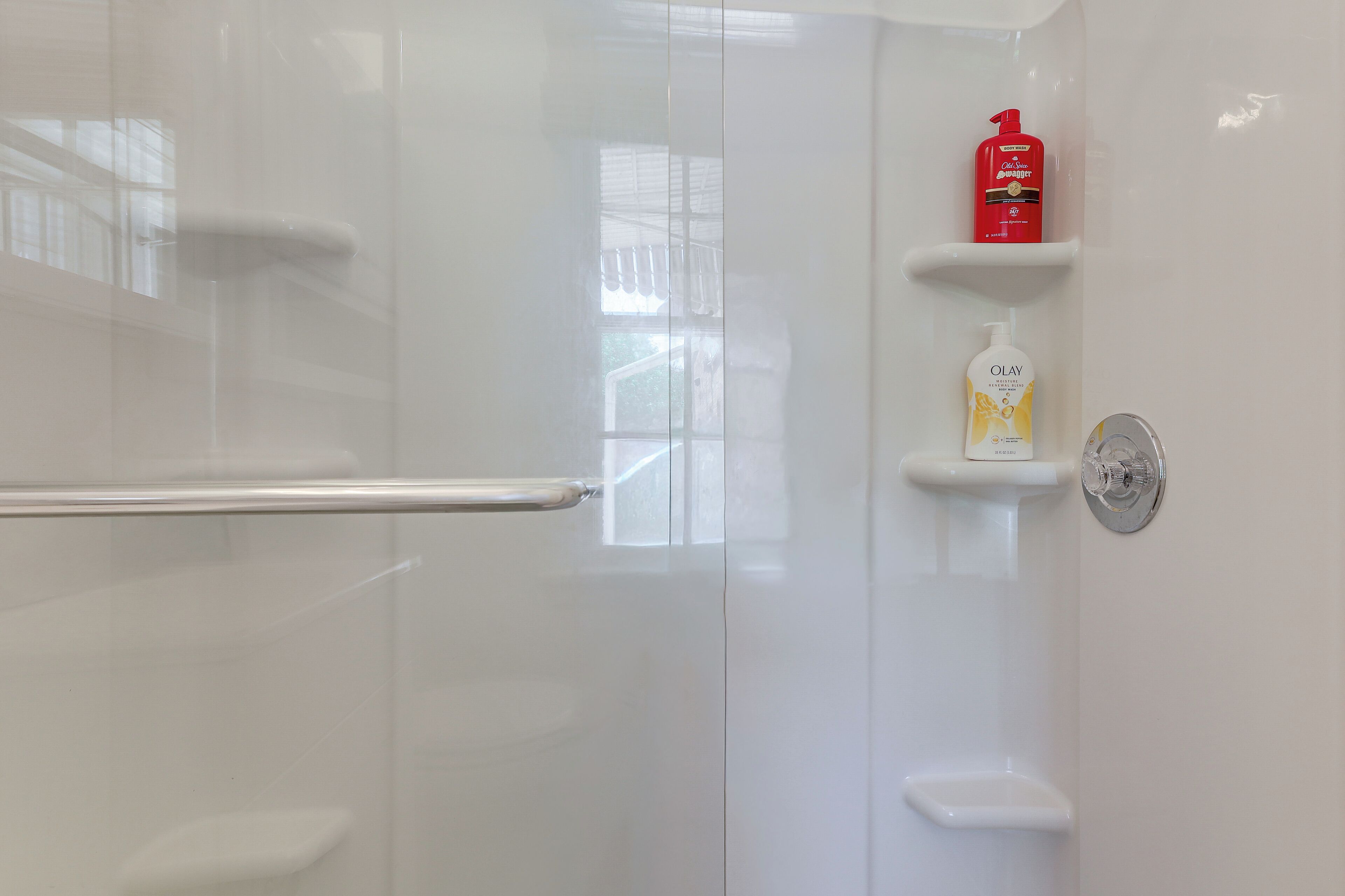 Une salle de bain avec une porte de douche en verre, une barre d’appui, un flacon de shampoing rouge et un flacon de lotion blanche posés sur une étagère.