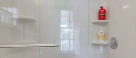 Une salle de bain avec une porte de douche en verre, une barre d’appui, un flacon de shampoing rouge et un flacon de lotion blanche posés sur une étagère.