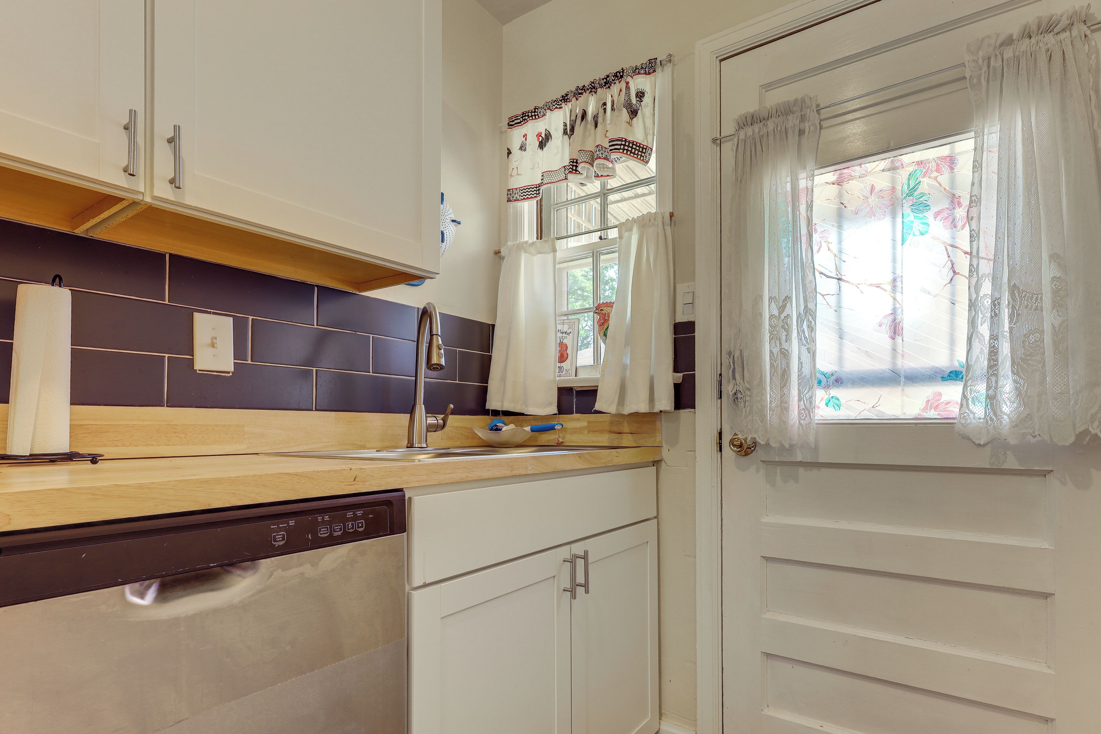 A kitchen with white cabinets, a dishwasher, a window with floral curtains, and a tiled backsplash.