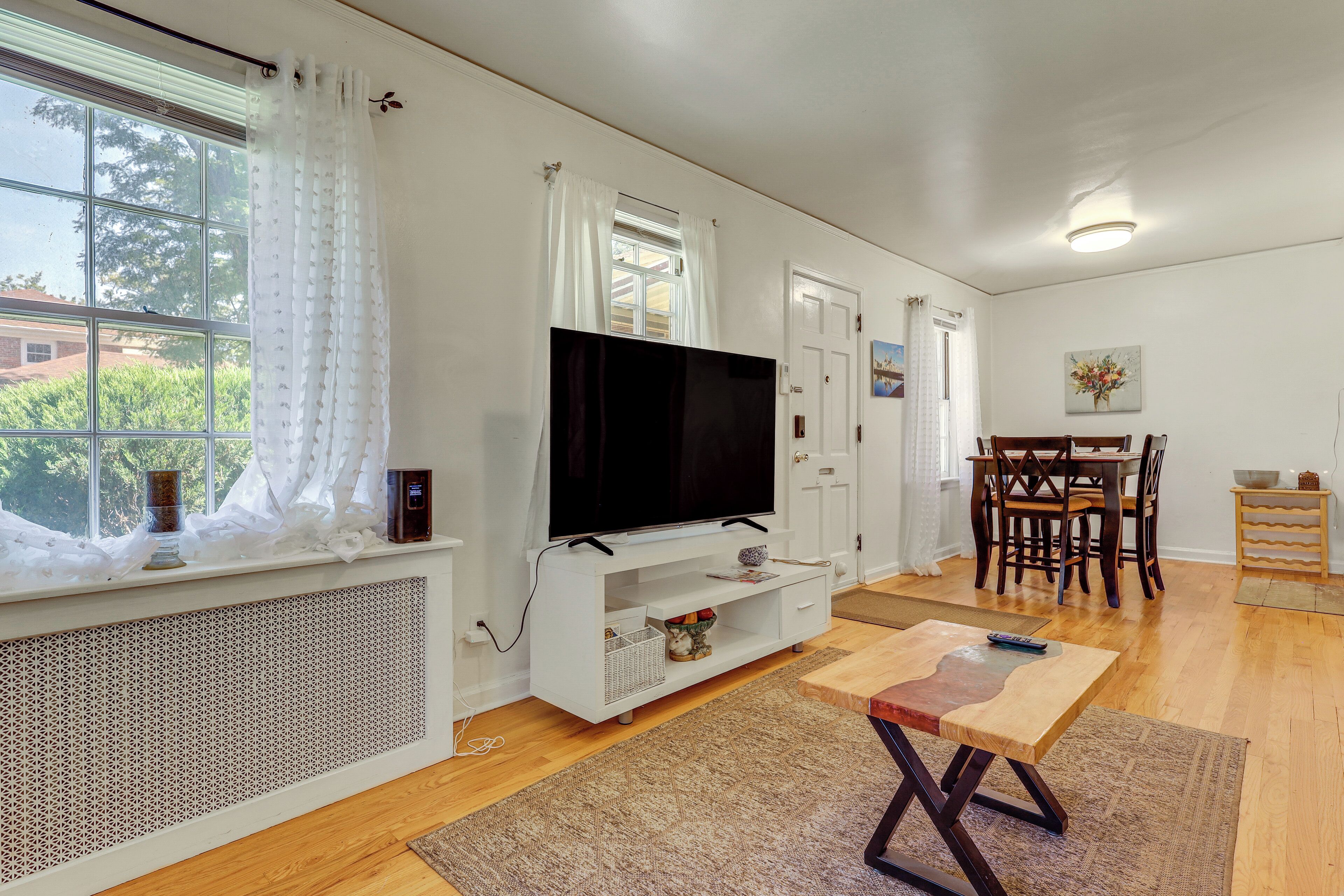 A modern living room with a flat-screen TV, a dining area with wooden furniture, and a large window with white curtains.