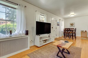 A modern living room with a flat-screen TV, a dining area with wooden furniture, and a large window with white curtains. - Nurse-friendly Rental Near Hospitals & Dtwn Pueblo (Pueblo)