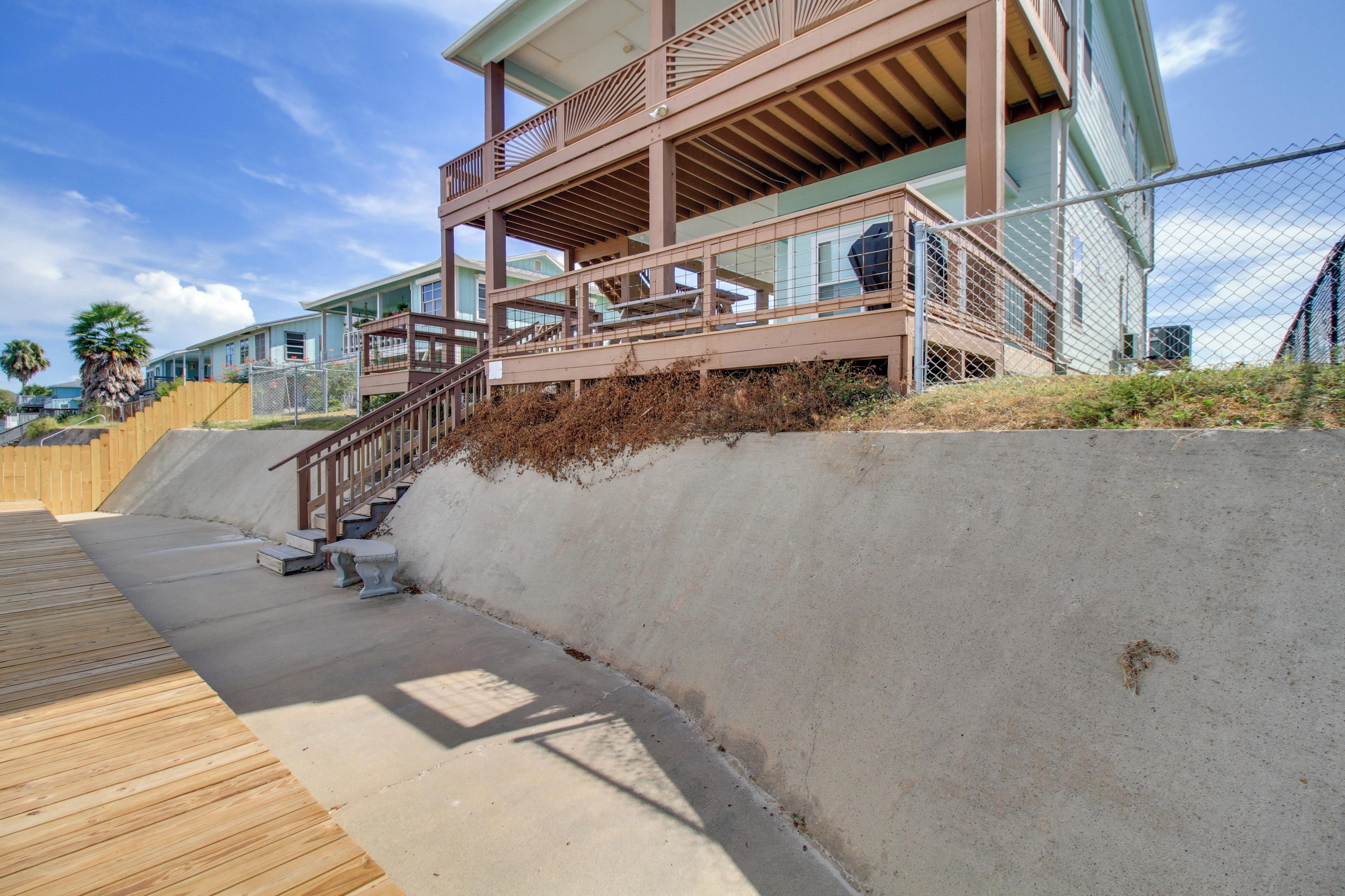 Een pand aan het strand met een houten terras, een betonnen steunmuur en een trap naar een balkon.
