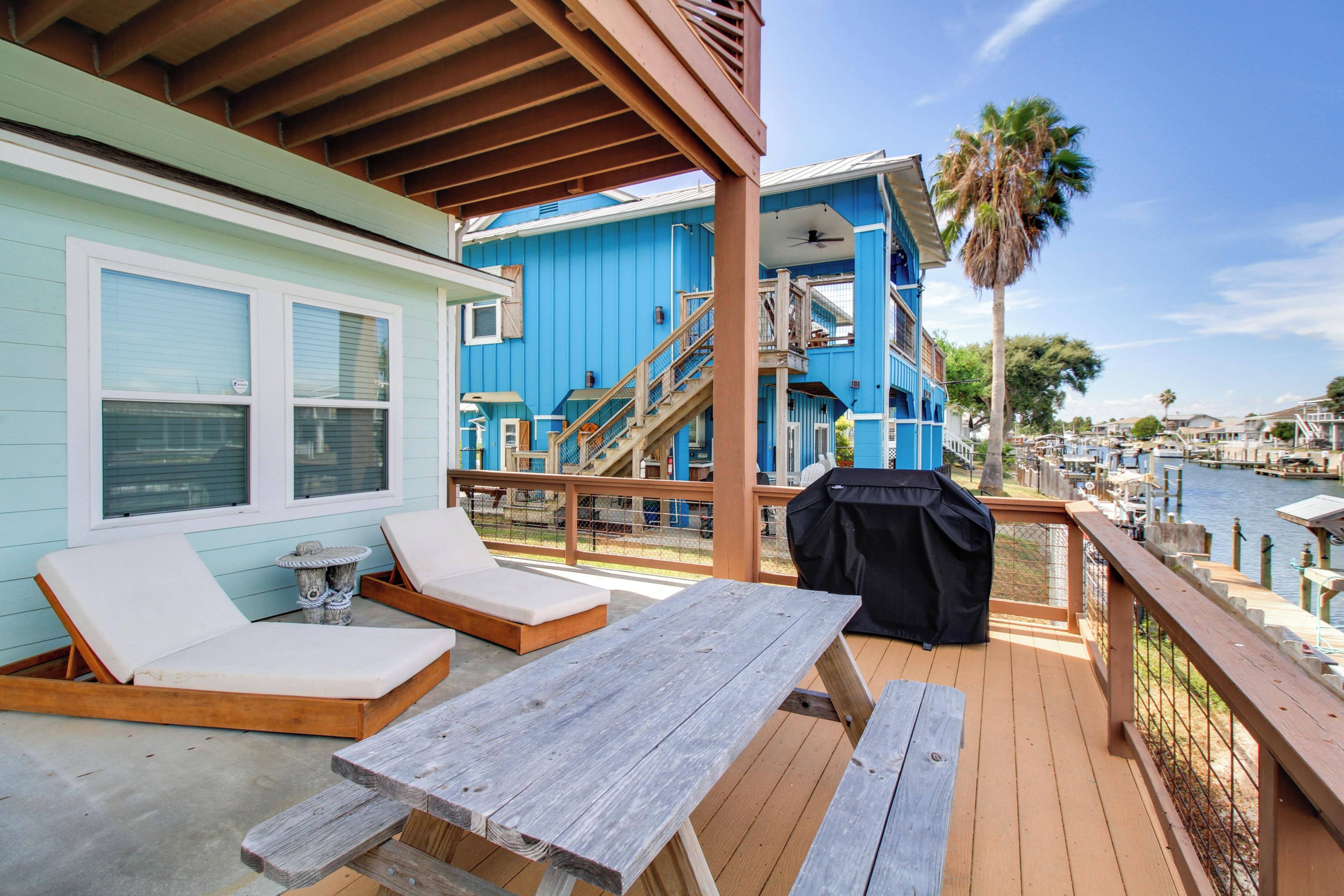 A wooden deck with a picnic table and lounge chairs, overlooking a marina with boats and a blue building with a staircase.