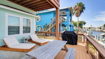 A wooden deck with a picnic table and lounge chairs, overlooking a marina with boats and a blue building with a staircase.