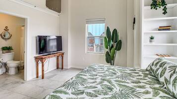 A bedroom with a bed, a television on a wooden table, a large plant, a window, and a bookshelf.