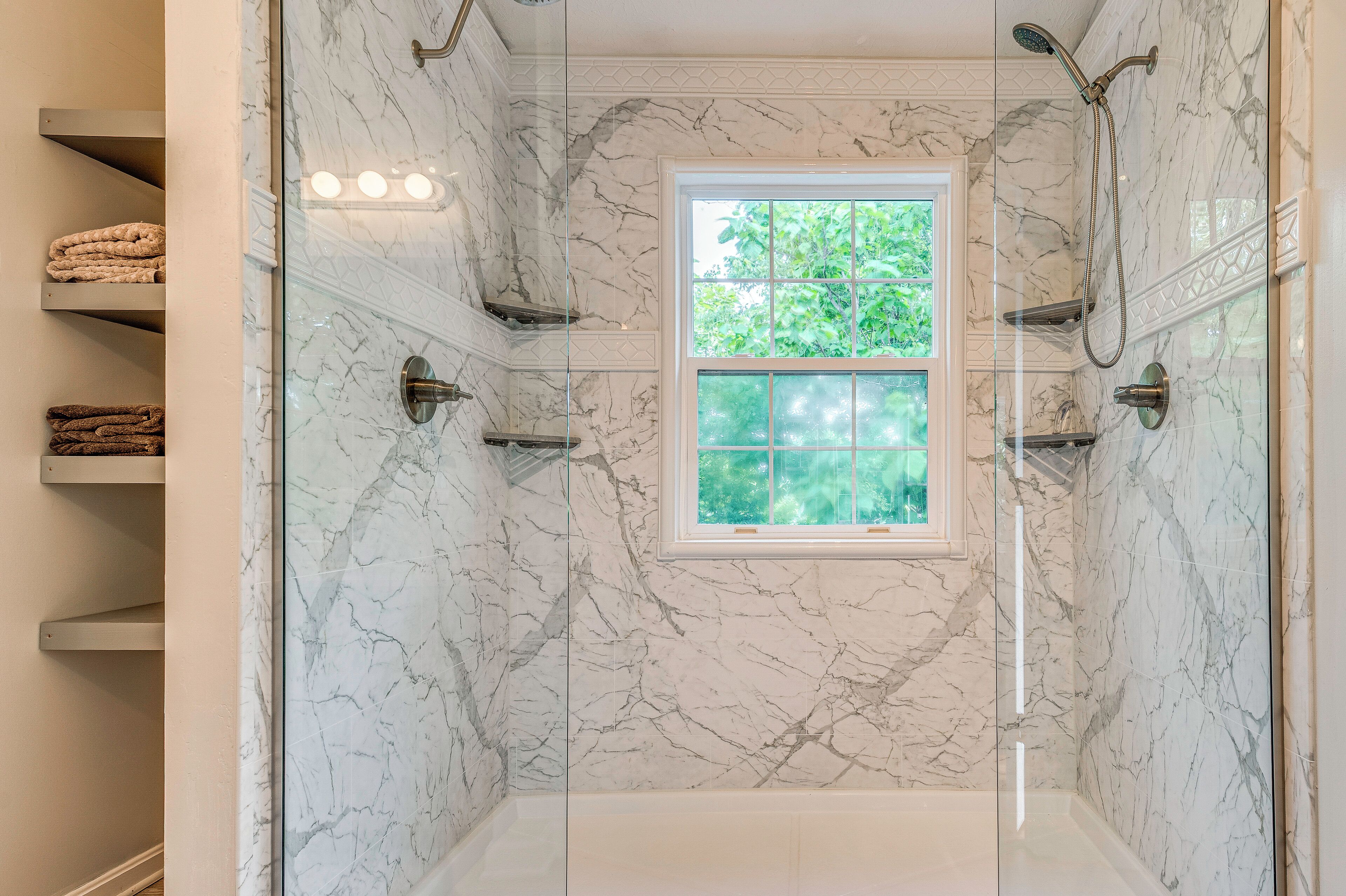 A bathroom with a glass shower enclosure, marble walls, a window, and towel racks.