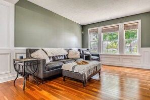 A living room with a dark leather sofa, a wooden coffee table, and a view of greenery through large windows. - Treehouse Views! Walking Distance to IMS (Indianapolis)